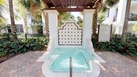 This outdoor spa area centers around a beautifully appointed hot tub, distinguished by its ornate tiled backsplash featuring a classic diamond-pattern design in white, brown, and multicolored accents. The decorative wall panel rises in an arched, architectural silhouette, framed by two stately white columns that support a wooden pergola overhead. The pergola provides partial shade while remaining open to the sky, creating a bright and airy atmosphere. Brick pavers form the surrounding deck, adding a warm, traditional character to the space. Lush tropical landscaping, including tall palm trees and dense foliage, encloses the area, offering a sense of privacy and a resort-like ambiance. A metal handrail provides convenient access into the spa. The clear, turquoise water of the hot tub contrasts attractively with the crisp white tiling and tilework surround. Signage visible within the space indicates posted spa rules, reflecting a well-maintained, regulated facility. Overall, the space combines refined architectural detailing with natural tropical elements to deliver a tranquil and visually appealing outdoor relaxation environment.
