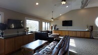 This single-floor space centers around a generously sized living area that exudes a warm and inviting ambiance. The room features vaulted ceilings that lend a sense of openness and volume, complemented by recessed lighting and dual ceiling fans that ensure comfort throughout. Large windows along one wall flood the interior with natural light while framing picturesque views of the surrounding greenery, creating a seamless connection to the outdoors. The living area is anchored by rich, dark leather sofas and ottomans arranged around an ornately carved wooden coffee table, offering both style and comfort. A wall-mounted flat-screen television is positioned prominently for easy viewing, backed by a run of warm wood cabinetry with dark countertops that provide generous storage and display space. The neutral-toned walls and carpeted flooring contribute to the room's relaxed, comfortable atmosphere. Decorative accents, including tall vases with dried botanicals and elegant table accessories, add refined touches throughout. The overall design balances casual comfort with polished detail, making this a welcoming and functional living space suited for both everyday relaxation and entertaining.