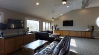 This single-floor space centers around a generously sized living area that exudes a warm and inviting ambiance. The room features vaulted ceilings that lend a sense of openness and volume, complemented by recessed lighting and dual ceiling fans that ensure comfort throughout. Large windows along one wall flood the interior with natural light while framing picturesque views of the surrounding greenery, creating a seamless connection to the outdoors. The living area is anchored by rich, dark leather sofas and ottomans arranged around an ornately carved wooden coffee table, offering both style and comfort. A wall-mounted flat-screen television is positioned prominently for easy viewing, backed by a run of warm wood cabinetry with dark countertops that provide generous storage and display space. The neutral-toned walls and carpeted flooring contribute to the room's relaxed, comfortable atmosphere. Decorative accents, including tall vases with dried botanicals and elegant table accessories, add refined touches throughout. The overall design balances casual comfort with polished detail, making this a welcoming and functional living space suited for both everyday relaxation and entertaining.