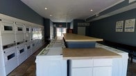 This single-floor laundry facility is designed with practicality and efficiency in mind, offering a clean and organized environment for laundry needs. The space is finished with deep navy-blue walls and a white ceiling with recessed lighting, giving it a crisp, modern appearance.

Along one wall, an extensive lineup of stacked front-loading washers and dryers provides ample capacity for multiple users. Each unit features a digital interface for ease of use. Centrally positioned throughout the room are generously sized white cabinetry units topped with light-toned laminate folding counters, offering plenty of surface area for sorting and folding laundry.

The floor is laid with durable stone-look tile, suitable for a high-traffic environment. Toward the rear of the space, natural light filters in through a glass-panel exit door, brightening the room alongside the recessed ceiling lights. A wall-mounted HVAC unit ensures year-round climate comfort for users.

Additional amenities visible in the space include a vending machine near the rear, a wall clock, and an emergency exit sign, all contributing to the facility's functional, user-focused design. The overall layout is open and spacious, allowing easy movement throughout the room while accommodating multiple users simultaneously.