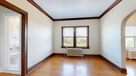 This single-floor residence reflects timeless character through a thoughtful blend of classic architectural details and functional living spaces. Throughout the unit, warm hardwood floors run continuously, complemented by rich dark wood trim and baseboards that frame each room with traditional elegance. A standout feature is the graceful arched doorway connecting the main living areas, adding a sense of openness and architectural distinction. A ceiling fan overhead enhances airflow and comfort in the living space. Natural light filters through double-hung windows fitted with blinds, offering views of surrounding greenery and the neighborhood streetscape. Cast-iron radiators beneath the windows provide efficient radiant heating, a hallmark of classic construction. The kitchen is equipped with a full-size refrigerator, an electric coil range, and stone-patterned tile flooring, offering a practical and compact cooking space. The bathroom retains its vintage charm with classic white hex tile flooring, a pedestal-style sink, a full soaking tub, and a frosted glass window for natural light and privacy. Interior doors feature original wood framing with glass panel insets, further highlighting the period craftsmanship. The layout flows naturally from room to room, creating a cohesive and characterful living environment.