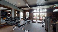 This fitness center combines industrial character with a fully outfitted workout environment, designed to accommodate a wide range of exercise routines. The space is anchored by striking architectural details, including exposed wooden ceiling beams, exposed brick accent walls, and large arched and multi-pane windows that flood the interior with natural light and views of the surrounding greenery. The main workout area houses a row of Cybex treadmills and elliptical machines positioned along the windowed wall, alongside a multi-station cable and weight machine for comprehensive strength training. A dedicated free weights section features a full dumbbell rack with floor-to-ceiling mirrors and an adjustable workout bench. A secondary area includes additional cardio equipment such as a rowing machine and stationary bike, set against a dramatic dark-painted accent wall adorned with a fitness-themed chalk art mural. A bright, separate zone offers a multi-functional cable machine, a colorful medicine ball rack, a punching bag, and a storage cabinet, with a posted workout board nearby. Recessed lighting and ceiling fans provide comfort throughout, while durable rubber flooring ensures safety and functionality across the entire space.