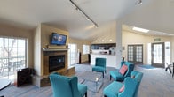 This single-floor space is thoughtfully designed to provide a comfortable and inviting environment for gathering and relaxation. The open-plan layout seamlessly connects the living area to the kitchen, creating a sense of spaciousness and flow throughout.

The living area is a standout feature, anchored by a tile-surround fireplace with a mounted flat-screen television above, serving as a natural focal point. Vaulted ceilings add to the airy atmosphere, while track lighting provides functional, adjustable illumination across the space. A set of vibrant teal upholstered armchairs with nail-head trim detailing surrounds a sleek glass coffee table, adding a pop of color and a modern aesthetic. Natural light filters in through multiple windows, enhancing the welcoming ambiance.

The kitchen is well-appointed with dark cabinetry, a breakfast bar-style counter, and modern appliances including a microwave and refrigerator. Pendant lights hang above the counter, providing warm task lighting and complementing the overall design.

The combination of vaulted ceilings, an open layout, a cozy fireplace, and contemporary furnishings makes this space functional yet stylish — well-suited for communal use or social gatherings.