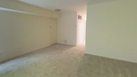 This single-floor residential space offers a clean, straightforward layout designed for practical everyday living. The main living area is finished with wall-to-wall carpet in a neutral beige tone, providing a soft underfoot feel that extends throughout the space. The walls are painted in a light cream and white palette, contributing to a bright, airy atmosphere even in the absence of abundant natural light. A flush-mount ceiling light fixture provides overhead illumination, keeping the space well-lit. A standard interior door is visible along one wall, offering access to an additional room or closet, while an open corridor leads further into the unit, suggesting a connected layout between living and private areas. A ceiling vent indicates a central HVAC system for climate control. The overall design is minimal and unadorned, emphasizing functionality and ease of use. The open feel of the main room provides flexibility for a variety of furniture arrangements, making it a versatile living area. The consistent flooring and color scheme throughout create a cohesive, unified aesthetic across the space.