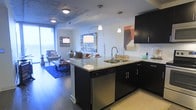 This single-floor space showcases a thoughtfully designed open-concept layout that seamlessly integrates the kitchen and living area. The kitchen is a standout feature, equipped with dark espresso cabinetry that contrasts elegantly against light granite countertops. A central island with a double undermount sink serves as both a functional workspace and a visual anchor for the space. Stainless steel appliances, including a dishwasher, complement the modern aesthetic, while white subway tile backsplash adds a clean, classic touch. Pendant lighting hangs above the island, providing focused task illumination alongside recessed ceiling fixtures. The exposed concrete ceiling lends an industrial character to the space. The open living area flows naturally from the kitchen, featuring hardwood-style flooring and large windows that invite natural light deep into the interior. Colorful artwork adorns the walls, adding visual interest and warmth. The overall ambiance balances contemporary urban design with comfortable livability, creating a cohesive and stylish environment well-suited for both entertaining and everyday living.