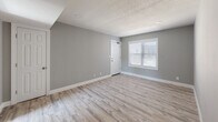 This single-floor space presents a clean, contemporary aesthetic defined by consistent light wood-look luxury vinyl plank flooring and soft gray walls with crisp white baseboards running throughout. The layout flows efficiently from the main living area toward the kitchen and additional rooms, creating an open, connected feel.

The living area is generously proportioned, featuring a textured ceiling, recessed lighting, and multiple HVAC vents ensuring comfortable climate control. Natural light enters through white-framed windows fitted with horizontal blinds, offering views of the surrounding neighborhood. A white paneled entry door anchors one end of the space.

The kitchen is a functional highlight, showcasing gray cabinetry, white subway tile backsplash, light countertops, and an electric range with an over-range microwave. A large window above the counter area floods the kitchen with abundant natural light, enhancing the bright, airy atmosphere.

Additional rooms continue the same neutral color palette and flooring, with windows providing outdoor views of neighboring rooftops and mature trees. The overall ambiance is modern, clean, and move-in ready, with cohesive finishes that lend the space a refreshed, well-maintained character throughout.