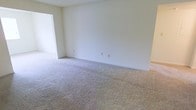 This single-floor space presents a clean, neutral aesthetic throughout, making it a versatile and welcoming environment. The interior is finished with soft, wall-to-wall carpeting in a light beige tone, providing a warm and comfortable feel underfoot. The walls are painted in a crisp, bright white, lending the space an open and airy atmosphere that maximizes the sense of light and volume.

The main living area is a well-proportioned open room with smooth white walls and standard electrical outlets conveniently positioned along the baseboard. A doorway opening to an adjoining room adds to the functional flow of the layout, suggesting a connected and accessible floor plan. The transition between rooms is clean and straightforward, with consistent flooring and wall finishes carrying throughout.

The overall ambiance of the space is minimal and neutral, providing an ideal blank canvas for a variety of uses and interior styles. The absence of clutter and the uniformity of finishes give the space a tidy, move-in ready quality. Whether used as a residential or professional setting, the layout and design offer practicality and adaptability in equal measure.