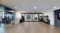 This single-floor fitness space is designed with both functionality and aesthetics in mind. The room features wide-plank hardwood flooring throughout, providing a clean and durable surface suited for a variety of exercises. Recessed lighting is evenly distributed across the white ceiling, ensuring bright, consistent illumination across the entire area.

A defining feature of the space is the expansive floor-to-ceiling mirror wall framed in black, which visually doubles the room's depth and allows users to monitor their form during workouts. To the right, a well-organized equipment rack holds an array of kettlebells, dumbbells, and free weights in ascending order, making them easily accessible. Additional equipment, including resistance tools and a punching bag, is positioned along the left wall, which also incorporates a textured glass partition that adds a stylish architectural detail.

The open central floor area provides ample room for stretching, functional training, or group exercise. The overall design balances a sleek, minimalist aesthetic with practical workout infrastructure, creating a professional-grade fitness environment.