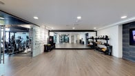 This single-floor fitness space is designed with both functionality and aesthetics in mind. The room features wide-plank hardwood flooring throughout, providing a clean and durable surface suited for a variety of exercises. Recessed lighting is evenly distributed across the white ceiling, ensuring bright, consistent illumination across the entire area.

A defining feature of the space is the expansive floor-to-ceiling mirror wall framed in black, which visually doubles the room's depth and allows users to monitor their form during workouts. To the right, a well-organized equipment rack holds an array of kettlebells, dumbbells, and free weights in ascending order, making them easily accessible. Additional equipment, including resistance tools and a punching bag, is positioned along the left wall, which also incorporates a textured glass partition that adds a stylish architectural detail.

The open central floor area provides ample room for stretching, functional training, or group exercise. The overall design balances a sleek, minimalist aesthetic with practical workout infrastructure, creating a professional-grade fitness environment.