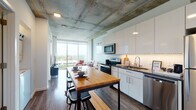 This contemporary urban unit showcases an industrial-modern aesthetic defined by dramatic exposed concrete ceilings and rich hardwood-style flooring throughout. The open-concept layout seamlessly connects the kitchen, dining, and living areas into a cohesive, functional space. The kitchen is outfitted with stainless steel appliances, crisp white cabinetry, a subway tile backsplash, and a butcher-block topped rolling island that doubles as a dining table with bench seating. The living room features a light neutral sofa paired with a vibrant red leather accent chair, exposed concrete columns, and soft blue-gray walls adorned with modern artwork. Floor-to-ceiling windows flood the living area with abundant natural light and frame sweeping elevated views of the surrounding cityscape. The bedroom continues the floor-to-ceiling window motif, offering panoramic city views, a bold teal accent wall, and the same exposed concrete ceiling that lends the unit its distinctive character. The bathroom is clean and well-appointed, featuring warm-toned tile flooring, a wood-framed vanity with an integrated sink, and a large mirror. Throughout the space, the interplay of raw industrial elements and refined modern finishes creates a sophisticated yet inviting urban living environment.