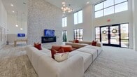 This contemporary amenity space is defined by its dramatic double-height ceilings, which create an expansive, airy atmosphere throughout. The main lounge area serves as the centerpiece, featuring a large sectional sofa arrangement in neutral tones accented with terracotta and burgundy pillows, anchored by a sleek wall-mounted fireplace set against a striking terrazzo-clad feature wall. A sculptural globe chandelier adds a modern touch overhead, while an abundance of natural light pours in through floor-to-ceiling black-framed windows that frame views of the surrounding neighborhood.

Adjacent to the lounge, a dedicated game room offers both a billiards table and shuffleboard tables, complemented by wall-mounted displays and curated framed artwork, all beneath the same soaring ceilings. The space transitions smoothly into a well-appointed lobby area, where warm wood-paneled walls, recessed lighting, and a collection of tasteful artwork create an inviting, gallery-like corridor.

Throughout the space, light-toned luxury flooring with a subtle abstract pattern unifies each zone seamlessly. The overall design balances clean, minimalist architecture with carefully chosen furnishings and art, resulting in a sophisticated yet approachable communal environment ideal for gathering, recreation, and relaxation.
