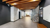 Located at 2150 Boulevard De Maisonneuve Ouest in Montréal, this building lobby offers a polished and well-appointed entrance experience. The space is defined by a striking combination of marble-clad walls — featuring both light, veined white marble and darker grey-toned marble panels — that lend an air of elegance throughout. Underfoot, large-format grey porcelain floor tiles provide a clean, contemporary foundation that complements the refined finishes above.

A standout architectural feature is the warm wood slat ceiling, with rich timber beams running across the length of the main corridor, adding texture and warmth that contrast beautifully with the darker ceiling accents and marble surfaces. Vertical wood slat partitions further echo this natural material palette, providing visual separation while maintaining an open feel.

The lobby comprises two distinct areas: an entry corridor leading from the glass-fronted street entrance — which allows natural light to filter through — and a more intimate inner zone housing the elevators, framed by dramatic dark marble surrounds. A cozy lounge seating area with accent furniture adds a residential warmth near the elevator bank.

A dedicated mailroom area features a full wall of individual metal mailboxes, marble-paneled walls, and a dark ceiling, providing residents with a functional yet cohesive space that aligns with the lobby's overall design language.