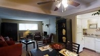 This single-story space offers an open and functional layout that seamlessly connects the dining and living areas. The dining area in the foreground is anchored by a dark wood table paired with matching chairs and decorative placemats, creating a welcoming setting for meals. A decorative bowl serves as a centerpiece, adding a touch of warmth to the arrangement.

The living area beyond features a white brick fireplace as a focal point, flanked by cushioned armchairs that invite relaxation. A low coffee table sits atop carpeted flooring, completing the seating arrangement. A tall black shelving unit along one wall provides both storage and display space, housing decorative vases and trailing greenery that add natural accents throughout the room.

Natural light filters in through a large window, brightening the space during the day, while a table lamp in the corner offers softer ambient lighting. Overhead, a ceiling fan with an integrated light fixture provides both air circulation and additional illumination. The overall ambiance is cozy and lived-in, with a neutral color palette and classic furnishings that lend the space a comfortable, homey character.