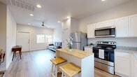 This thoughtfully designed single-story home offers an efficient and comfortable layout across one floor. The open-concept living and kitchen area forms the heart of the home, featuring light wood-look flooring, recessed lighting, and a ceiling fan that flows seamlessly from one space to the next. The kitchen is well-appointed with white shaker cabinetry, granite countertops, a full suite of stainless steel appliances including a refrigerator, range, microwave, and dishwasher, and a central island with a butcher block countertop and bar seating — ideal for casual dining or entertaining. The living room is bright and airy, with a large window overlooking the outdoors and a ceiling fan overhead. A welcoming entryway with clean lines and natural light provides a smooth transition into the home. The bedroom offers a peaceful retreat with multiple windows, a ceiling fan, and warm neutral tones, complemented by plush area rugging and wood-look flooring. The bathroom is clean and modern, featuring a wide vanity with white cabinetry, a large mirror, and a natural light window. A dedicated closet, two hallways, and two balconies round out this functional and inviting home, with an outdoor area featuring a small lawn and gravel yard visible from the exterior.