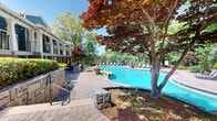 Located at 1701 Dawsonville Hwy in Gainesville, Georgia, this expansive outdoor amenity space offers a resort-caliber experience designed for relaxation and recreation. At its centerpiece sits a generously sized freeform swimming pool with vivid blue water, complemented by a natural stone water feature that adds both visual appeal and a soothing ambiance. Adjacent to the main pool is a circular hot tub, providing a dedicated space for leisure. The pool deck is finished in classic brick pavers that extend throughout the entire outdoor area, creating a cohesive and durable surface underfoot. Terraced stone retaining walls, constructed from stacked natural stone, add architectural dimension to the landscape while integrating seamlessly with the surrounding greenery. Mature trees — including striking Japanese maples with deep red foliage — and lush shrubs provide shade and privacy throughout the space. An outdoor kitchen station, framed in matching stone masonry, offers a built-in grill and prep area ideal for entertaining. A covered colonnaded structure with brick columns and an upper-level balcony borders the space, providing additional shade and direct interior access. Lounge chairs and patio seating are positioned throughout, inviting guests to enjoy the open-air setting beneath the Georgia sky.