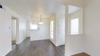 This single-floor residence offers a clean, open layout designed for comfortable everyday living. The entryway greets visitors with a solid white paneled front door flanked by windows fitted with blinds, allowing natural light to filter throughout the space. Dark hardwood flooring runs through the main living and dining areas, providing a rich contrast against the crisp white walls and trim. A pendant light fixture hangs above the dining area, adding a focused decorative element that anchors the space. Structural columns subtly define zones within the open floor plan without compromising the airy, connected feel of the interior. The kitchen is visible beyond the dining area and features white cabinetry and a pass-through or counter window that enhances the sense of openness. Additional windows throughout the space ensure consistent natural light from multiple directions, contributing to the overall brightness and welcoming ambiance. The neutral color palette of whites and warm wood tones creates a versatile backdrop well-suited to a variety of interior styles. The layout flows intuitively, with each area transitioning smoothly into the next, making the home feel both functional and inviting.
