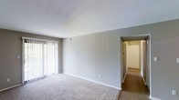 This single-floor space presents a straightforward and functional layout with a focus on clean, neutral aesthetics. The main living area features wall-to-wall carpeting in a light beige tone, contributing to a soft and comfortable atmosphere. Walls are finished in a subtle gray-white palette, complementing the overall understated design. A large window fitted with vertical blinds dominates one wall, allowing for adjustable natural light and a bright, open feel. A ceiling-mounted HVAC vent indicates a centralized climate control system, ensuring year-round comfort. The living area transitions seamlessly into a hallway visible toward the back of the space, which features hard-surface flooring — likely wood or laminate — providing a practical contrast to the carpeted main area. The hallway leads to what appears to be additional rooms, including a door that may access a closet or entry point. White baseboards run throughout, adding a crisp, finished detail to the space. Overall, the interior offers a clean, move-in-ready environment with a neutral canvas well-suited for a variety of furnishing styles and personal tastes.