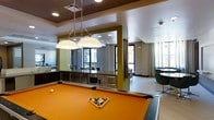 This single-floor communal space is designed to serve multiple functions within an open, well-appointed layout. The focal point of the room is a full-size pool table with a vibrant orange felt surface, positioned beneath a pair of dome-shaped pendant lights that provide direct, focused illumination. The ceiling features a combination of recessed lighting and a contrasting tray design with warm brown tones, adding architectural interest overhead.

Adjacent to the billiards area, a built-in kitchenette offers dark cabinetry, stainless steel appliances including a microwave and warming drawer, and a sleek countertop — ideal for entertaining. A large mirror panel positioned near the kitchenette visually expands the space and reflects natural light from the surrounding windows.

To the side, a lounge area features modern seating with dark round tables and minimalist chairs, bathed in daylight from floor-to-ceiling windows dressed with sheer curtains. The flooring transitions between tile and other surfaces, delineating functional zones. Overall, the space presents a contemporary, multi-use amenity environment suited for social gatherings and recreation.