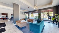 Located on Church Street in Glenview, Illinois, this single-floor community lounge space offers a thoughtfully designed interior that blends contemporary style with comfortable, functional living areas. The space is organized into a living room and dining room, connected by open passageways that promote a seamless flow throughout.

The living area serves as the centerpiece of the space, anchored by a sleek, linear stone fireplace set against a white stacked-stone accent wall. Tray ceilings add architectural depth overhead, while cluster pendant lights with amber-toned bulbs cast a warm glow. Seating includes a bold teal velvet sofa and tufted caramel leather chairs, arranged atop a navy area rug over light-toned tile flooring. Large floor-to-ceiling windows flood the room with natural light and offer views of the surrounding streetscape.

A secondary seating nook features a vibrant blue wingback chair positioned before a textured geometric accent wall adorned with decorative wall-mounted deer heads and an inset display screen, adding a playful and eclectic design element.

The dining area provides a bright, open setting with a modern black table and upholstered gray chairs beneath soaring ceilings. Oversized windows frame lush potted plants and allow daylight to pour in from multiple angles. Adjacent rooms, visible through open doorways, hint at additional amenity spaces including a kitchen bar and lounge area, rounding out this well-appointed communal retreat.