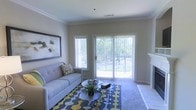 This single-floor space showcases a well-appointed living room designed with comfort and functionality in mind. The room features neutral beige walls accented by elegant crown molding, creating a polished and cohesive aesthetic. Natural light floods the space through a combination of a side window fitted with blinds and a large sliding glass door that opens to an outdoor area, offering pleasant views of surrounding greenery. The sliding door serves as a focal point of the room, framing the outdoor scenery while enhancing the sense of openness and airiness within. The living area is finished with plush carpeting and anchored by a bold geometric-patterned area rug in shades of gray, yellow, and white, adding a contemporary touch. A tufted gray sofa paired with a round coffee table arrangement provides a practical yet stylish seating area. Wall décor includes a sizable abstract framed artwork, contributing to the modern character of the space. A fireplace surround is partially visible along one wall, suggesting an additional cozy feature. The overall ambiance is light, airy, and inviting.