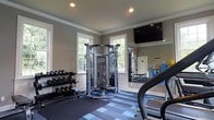 Located at 1390 Christopher Wren Dr in Gahanna, Ohio, this fitness center offers a well-equipped, single-floor workout facility designed for a comprehensive exercise experience. The space features elegant crown molding, white wainscoting, and neutral gray walls that create a clean, polished aesthetic. Large windows on multiple walls flood the room with natural light, contributing to an open and energizing atmosphere. The flooring consists of a patterned blue and gray modular carpet tile, providing both comfort and durability underfoot. At the center of the room stands a multi-functional cable machine, ideal for a wide range of strength training exercises. A full dumbbell rack lines one wall, offering a complete set of free weights for varied workouts. Cardio equipment, including a treadmill, is positioned for easy access. A large wall-mounted mirror expands the sense of space while allowing users to monitor their form. A wall-mounted flat-screen television adds an entertainment element to workouts. Recessed ceiling lighting ensures the space is well-lit throughout. The overall ambiance is professional and inviting, balancing functional fitness infrastructure with thoughtful architectural details.