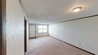 This single-floor residence offers a practical and functional layout with a variety of well-defined living spaces throughout. The generous living area is finished with plush wall-to-wall carpeting, neutral-toned walls, dark wood baseboards, and a textured ceiling, with a large front-facing window and direct access to the entry door, allowing natural light to fill the room. The kitchen is equipped with rich dark wood cabinetry providing ample upper and lower storage, a double-basin sink with dark countertops, a white electric coil-burner range, and a built-in dishwasher, all set over durable wood-look flooring. The bedrooms are carpeted and generously sized, each featuring windows with blinds that bring in natural light. One bedroom includes a built-in low-profile wood cabinet unit spanning the length of one wall, offering additional storage and display space. The full bathroom features a single-sink vanity with a marble-patterned countertop, a wall-mounted mirror with overhead lighting, a toilet, and a full tub-shower combination, complemented by wood-look flooring. A dedicated laundry room houses a full-size washer and dryer alongside a water heater, with wood-look flooring and neutral walls.