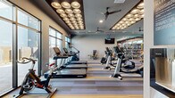 Located at 3600 W Happy Valley Rd in Phoenix, AZ, this well-appointed fitness center spans a generous single floor divided into two distinct areas designed to support a comprehensive workout experience. The main gym area is expansive and thoughtfully equipped with a full lineup of Life Fitness cardio machines, including treadmills, ellipticals, and stair climbers, alongside a robust selection of strength training equipment such as cable machines, weight benches, and a well-stocked dumbbell rack. A bold, dynamic mural serves as a striking focal point on one wall, adding energy and visual interest to the space. The ceiling features a signature cluster of dome-shaped pendant lights set within a coffered panel, creating a modern and polished ambiance, while multiple ceiling fans ensure air circulation throughout. Floor-to-ceiling windows along multiple walls flood the space with natural light and offer views of the surrounding outdoor areas. The flooring combines striped carpet tiles in the main zone with hardwood-style flooring in the secondary room. The dedicated cycling and cross-training studio houses spin bikes, a rowing machine, stability equipment, and a large wall-mounted television, making it ideal for group or individual training sessions. Black-framed glass doors connect the two areas seamlessly, reinforcing the facility's contemporary, cohesive design aesthetic.