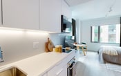 This thoughtfully designed single-floor studio delivers a clean, modern aesthetic throughout its compact yet well-organized layout. The galley-style kitchen runs along one wall and is outfitted with handleless white cabinetry extending floor to ceiling, under-cabinet LED lighting, a white composite countertop, a brushed gold undermount sink, and integrated appliances including a built-in oven and microwave — maximizing both function and visual clarity. Light wood flooring flows continuously through the space, reinforcing a sense of cohesion and warmth against the predominantly white interior. The living and sleeping area is openly connected to the kitchen, creating a flexible, multi-purpose zone. A natural wood headboard panel anchors the sleeping area, complemented by open shelving and soft, neutral furnishings. A window with full-length dark curtains frames views of the surrounding urban streetscape, allowing natural light to filter in while providing privacy when desired. A small dining nook with a round table and teal accent chairs adds a functional social corner. The bathroom features pale blue ceramic tile flooring, soft grey walls, and a glass-enclosed shower with clean white paneling, maintaining the minimal and fresh character found throughout the rest of the space.