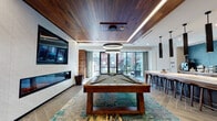 Located at 8320 University Executive Park Drive in Charlotte, NC, this single-floor amenity space showcases a cohesive contemporary design defined by rich wood-paneled ceilings and accent walls, large-format tile flooring in herringbone patterns, and expansive floor-to-ceiling windows that invite abundant natural light and outdoor views. The open layout connects several distinct zones that flow seamlessly into one another.

The lounge area features a teal sectional sofa, neutral accent seating, and a wall-mounted fireplace, anchored beneath dramatic pendant lighting with dark drum shades. The warm wood ceiling planks extend throughout, creating a unified and inviting atmosphere.

Adjacent to the lounge, a dedicated billiards area centers around a substantial wood-framed pool table beneath a circular ring chandelier, with a wall-mounted television and the same fireplace feature visible from multiple angles.

The kitchen and bar zone is highlighted by striking teal chevron-tiled walls, warm wood cabinetry, white quartz countertops, and a long island bar lined with wooden stools. Sleek pendant lights descend from above, while a coffee station and full cabinetry complete the space.

A corridor with herringbone tile flooring, wood accent walls, and glass-paneled doors provides access to additional private areas, reinforcing the sophisticated and functional character of the overall space.