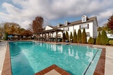 Located at 575 McDonough Pkwy in McDonough, Georgia, this outdoor amenity area serves as a central gathering space within a well-established residential community. The centerpiece is a generously sized swimming pool with clear, turquoise water and brick-trimmed edges, offering designated shallow and deeper sections clearly marked for safety. Surrounding the pool is a broad concrete deck outfitted with blue lounge chairs and umbrella-shaded seating areas, providing ample space for relaxation and recreation. A substantial wooden pergola runs along one side of the pool deck, offering shaded lounging beneath its open-beam structure and adding an architectural focal point to the outdoor space. On the opposite side of the pool, tiered landscaping with additional umbrella-shaded seating creates a layered, resort-like atmosphere. A smaller clubhouse or pool house structure with white siding, a covered portico supported by columns, and French doors is situated adjacent to the pool area, complemented by decorative plantings including columnar evergreens, ornamental trees, and seasonal foliage. The surrounding residential buildings feature classic white siding, dark shutters, and dormer windows, framing the amenity area with a cohesive architectural character. The entire space is enclosed by iron fencing and benefits from an open sky view, creating a bright and inviting outdoor environment.