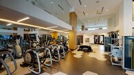 This fitness center at 480 K St SW in Washington, DC, is a thoughtfully designed, single-floor facility that blends contemporary aesthetics with comprehensive workout amenities. The space is defined by soaring double-height ceilings that create an open, airy atmosphere, enhanced by abundant natural light streaming through floor-to-ceiling windows that frame views of the surrounding urban landscape.

The flooring features a striking geometric hexagonal pattern in neutral tones, providing visual interest while delineating different workout zones throughout the space. Warm wood-clad structural columns punctuate the facility, adding an organic contrast to the otherwise clean, white architectural palette. Large wall mirrors amplify the sense of openness and allow users to monitor their form during exercise.

The cardio zone is well-appointed with a comprehensive selection of Matrix-brand treadmills, ellipticals, stair climbers, and stationary bikes, all equipped with individual digital displays. A dedicated strength training area houses a range of selectorized cable and resistance machines, while the open functional training zone accommodates performance equipment including battle ropes and a heavy training tire. Ceiling fans and recessed LED linear lighting contribute to a comfortable and well-illuminated workout environment throughout.