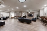 Located at 847 Development Drive in Kingston, Ontario, this single-floor amenity space offers a generous and thoughtfully designed communal environment ideal for socializing and recreation. The open-concept layout is anchored by light-toned wood-look flooring that flows seamlessly throughout, contributing to a bright and cohesive aesthetic.

The main lounge area is a standout feature, appointed with multiple black leather sofas and armchairs arranged around round coffee tables, creating inviting conversation zones. A wood-panelled accent wall serves as the backdrop for a large flat-screen television, accompanied by a decorative oversized clock and a console table — adding character and a contemporary-industrial flair. Pendant lighting fixtures hang from the ceiling, complementing the recessed pot lights that illuminate the space warmly.

A bar-height counter with leather-upholstered stools provides an additional casual seating area, while a dedicated dining zone accommodates group gatherings with long tables and industrial-style metal chairs. A separate, more intimate game room offers additional flexible table seating for activities or meetings.

Transitional areas feature framed wall art, a wall mirror, and decorative accents that enhance the polished, modern ambiance. Structural columns with wood-panel cladding add architectural interest, and the overall neutral colour palette of whites, greys, and warm wood tones creates a welcoming and versatile communal retreat.