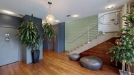 The lobby of The Pines Apartment Homes offers a well-designed and welcoming communal entrance space that balances modern aesthetics with natural warmth. The flooring throughout is warm-toned hardwood, providing a grounding contrast to the cool blue-gray walls that define the corridor and seating areas. A standout architectural feature is the eye-catching honeycomb-patterned mosaic tile accent wall in soft sage green, which lines the staircase and serves as a focal point of the interior. The staircase itself features clean concrete treads paired with a sleek white metal railing, contributing to the space's contemporary character. Organic-shaped stone or resin ottomans are arranged near the base of the stairs, offering casual seating with a sculptural quality. The entry area near the street-facing facade benefits from abundant natural light through large windows, with views of mature trees outside. A branded entry mat and a simple bench near the front provide functional touches. Large potted tropical plants are placed throughout, adding lush greenery that softens the overall aesthetic. Recessed lighting and a globe pendant fixture provide ambient illumination, while a mailbox alcove and corridor leading further into the building reinforce the space's practical, resident-focused design.