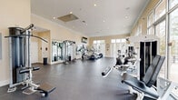 Located at 1904 East Palomar Street in Chula Vista, CA, this fitness center offers a generously sized, single-floor exercise space designed to support a comprehensive workout experience. The room features warm beige walls accented by elegant crown molding, recessed lighting throughout, and a high ceiling that contributes to the open, airy atmosphere. Large windows along multiple walls allow abundant natural light to fill the space while providing views of the surrounding outdoor environment, creating an inviting and energizing environment for exercise. The durable dark rubber flooring provides both safety and functionality across the entire workout area. Cardio equipment includes multiple treadmills with individual monitors, elliptical trainers, and a recumbent bike, while strength training is well-represented with a multi-station cable machine, various weight benches, and additional resistance equipment from brands such as Precor. Full-length mirrors along one wall enhance the sense of space and allow users to monitor their form during workouts. A wall-mounted flat-screen television is positioned above the mirror bank for entertainment. Glass entry doors provide easy access directly from the exterior, and the open floor plan allows for efficient equipment arrangement with ample circulation space between stations.
