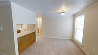 This single-floor residence offers a practical and comfortable layout designed for ease of living. The main living area is finished with warm beige carpeting that extends throughout, creating a soft and cohesive atmosphere. Crown molding runs along the perimeter of the ceiling, adding a refined architectural detail to the space, while a textured popcorn ceiling and a flush-mount ceiling fixture provide a classic residential feel.

A raised pass-through counter with wood-panel facing separates the kitchen from the living area, allowing for an open and connected flow between the two spaces. A glimpse into the kitchen reveals light cabinetry and standard appliances. A hallway visible from the living room leads to additional rooms, including what appears to be a bathroom with warm wood-toned cabinetry.

Natural light enters the living area through a window fitted with blinds, brightening the neutral, cream-toned walls. A floor-level air vent is built into the baseboard, indicating a central HVAC system. White baseboards frame the room cleanly throughout. The overall ambiance is understated and functional, with a neutral palette that provides a versatile backdrop suitable for a variety of interior styles.