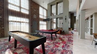 This expansive lounge is defined by its dramatic double-height ceilings and floor-to-ceiling windows that flood the interior with natural light. Exposed brick walls running throughout lend an industrial character, while herringbone-patterned tile accents and textured wall panels introduce refined, contemporary detailing. Bold, patchwork-style area rugs in deep reds and blues anchor the various zones within the space, providing visual continuity across the open layout.

A dedicated game area houses a full-size billiards table and a shuffleboard table, making it an ideal setting for social gatherings. Adjacent to this is a generous lounge zone furnished with a tufted leather sofa, upholstered armchairs, and a rocking chair arranged around a coffee table. A long bar counter lined with leather barstools runs alongside the window wall, offering additional seating with abundant natural light.

A more intimate lounge nook features a curved sectional sofa, tufted velvet wingback chairs, and decorative wall sconces, creating a cozy retreat within the larger space. The kitchen area includes stainless steel appliances, ample counter space, and a solid wood dining table beneath pendant lighting, all set against a warm exposed brick backdrop.