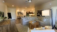 This single-floor residence offers a thoughtfully designed open-concept layout that integrates the living, dining, and kitchen areas into one cohesive, functional space. The living area is anchored by a generously sized sectional sofa in a soft neutral tone, complemented by a textured area rug and a rustic wooden coffee table that adds a natural, organic element to the room. Recessed lighting and a ceiling fan provide both ambiance and practicality, casting a warm, welcoming glow throughout the space. Hardwood-style flooring runs throughout, adding warmth and visual continuity. Decorative wall art featuring bold black-and-white photography lends an urban, contemporary character to the interior. The dining area sits adjacent to the living space, offering a seamless transition for entertaining. Beyond it, the kitchen features dark cabinetry, pendant lighting, and a breakfast bar with seating, making it both stylish and highly functional. The open layout encourages effortless flow between spaces, making this residence ideal for both everyday living and hosting guests. The overall ambiance is modern yet comfortable, with a neutral color palette and carefully chosen furnishings that create a balanced, welcoming atmosphere throughout.