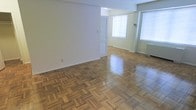 This single-floor space showcases a spacious, open layout highlighted by beautifully polished parquet hardwood flooring throughout. The intricate geometric pattern of the parquet adds a timeless, classic character to the interior. The main living area is generously proportioned, with clean white walls that complement the warm tones of the wood flooring.

Natural light is a standout feature of this space, with multiple large windows fitted with horizontal blinds allowing ample daylight to filter in. The windows contribute to a bright, airy atmosphere that enhances the sense of openness. A radiator unit is visible along one wall, indicating a traditional heating system.

The layout appears to extend into a secondary area or adjoining room, partially visible through a doorway, suggesting a functional and versatile floor plan. The transition between spaces is seamless, maintaining consistent flooring throughout. The overall ambiance is clean, neutral, and well-lit, providing a blank canvas that would suit a variety of interior styles and uses. The combination of classic architectural details and abundant natural light gives this space a welcoming and practical character.
