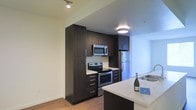 This modern single-floor residence is designed with a clean, contemporary aesthetic that emphasizes both functionality and style. The kitchen serves as a focal point of the space, showcasing rich dark espresso cabinetry that extends floor to ceiling, providing generous storage capacity. A suite of stainless steel appliances — including an electric range, over-the-range microwave, and refrigerator — adds a polished, professional look. Light-toned countertops offer a striking contrast against the dark cabinetry, while a kitchen island or peninsula extends the prep and dining area, making the space ideal for both everyday cooking and entertaining. A subtle tile backsplash adds texture and visual interest behind the range. The flooring features warm wood-tone hardwood or laminate planks that run throughout, lending a sense of continuity and warmth to the space. Recessed and flush-mount ceiling lighting ensures the space is well-illuminated, with additional track lighting enhancing the kitchen area. The open layout allows for a natural flow between the kitchen and adjoining areas, creating an inviting and practical living environment suited to modern lifestyles.