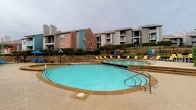 This residential community centers around a generously sized freeform swimming pool with clear turquoise water and a decorative fountain feature at its heart. The pool deck is finished in textured concrete and provides ample space for outdoor lounging, furnished with bright yellow and blue cushioned chairs and shade umbrellas positioned throughout. A raised sun terrace bordered by a rustic stacked-stone retaining wall offers an elevated lounging area with additional seating, overlooking the main pool below.

The surrounding landscape is carefully maintained, featuring trimmed hedges, flowering shrubs, and greenery that add a sense of privacy and visual appeal to the outdoor environment. Classic-style lamp posts are placed throughout the pool area, contributing to the community's character and providing ambient lighting.

The multi-story residential buildings surrounding the pool feature a mix of brick and stucco exteriors accented with teal and gray siding, with private balconies overlooking the common area. Secured black iron fencing and gated entry points are present at ground-level access areas, reinforcing the community's attention to safety and controlled access. The overall ambiance of the outdoor space is relaxed and resort-like, designed to serve as a central gathering and recreation hub for residents.