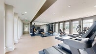 Located at 4901 Meridian Way in Frederick, Maryland, this fitness center is a thoughtfully designed, single-floor facility offering a comprehensive workout experience across two dedicated exercise rooms connected by a hallway. The space is finished with light hardwood-style flooring, crisp white crown molding, and neutral gray walls that create a clean, modern aesthetic throughout.

The main exercise room is expansive and light-filled, featuring a dramatic wall of large, grid-paned windows that frame views of an outdoor pool and patio area. Recessed lighting complements the abundant natural light, and the room is outfitted with a full suite of cardio and strength equipment, including stationary bikes, cable machines, rowing machines, and weight benches, all positioned on protective floor mats. A row of elegant white columns along the window wall adds an architectural detail that elevates the space.

The second exercise room offers a more intimate setting, ideal for functional training and flexibility work. It features the same light flooring and gray walls, along with a smart fitness mirror, a rack of medicine balls, and ample open floor space for stretching or mat-based workouts. Natural light filters in through large windows overlooking the landscaped grounds. Together, the two rooms provide a well-rounded, welcoming environment suited for a full range of fitness activities.