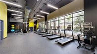 Located at 932 E 26th St in Hialeah, FL, this modern fitness center offers a generously sized, single-floor open layout designed to accommodate a full range of workout activities. The space features a striking industrial-inspired aesthetic, with an exposed dark ceiling showcasing visible ductwork and sleek linear LED lighting that illuminates the entire floor evenly.

Bold yellow accent walls contrast sharply against the dark charcoal and gray tones throughout, creating an energetic and motivating atmosphere. Durable rubber flooring covers the entire space, providing both safety and comfort underfoot.

The cardio zone lines the floor-to-ceiling glass window wall, where a row of treadmills and elliptical machines face lush outdoor views of palm trees and greenery, allowing natural light to flood the interior. Adjacent to the cardio area is a well-equipped strength training section featuring cable machines, a Smith machine, and a comprehensive dumbbell rack. A dedicated functional fitness area includes stability balls, resistance equipment, and open floor space for stretching or bodyweight exercises. Full-length mirrors are strategically placed to assist with form and amplify the sense of space. The facility also benefits from excellent ventilation provided by the industrial HVAC system overhead.