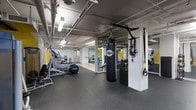 This fitness center offers a comprehensive workout environment designed to support a wide range of exercise routines. The space features an open, industrial-style layout with exposed ceiling pipes and overhead fluorescent lighting that contribute to a functional, no-frills atmosphere. Rubber flooring throughout the gym provides cushioning and traction for safe, high-impact training.

The cardio section is equipped with multiple machines, including an elliptical trainer and additional aerobic equipment, positioned to make the most of the available floor space. A TKO heavy punching bag hangs from a ceiling-mounted bracket alongside a pull-up bar and TRX suspension training straps, creating a dedicated functional fitness and boxing zone. Mirrored wall panels are strategically placed to allow users to monitor their form during exercise.

Structural columns divide the space into distinct workout zones while maintaining an open feel. Yellow accent walls add a bold pop of color that energizes the environment. Additional strength training benches and equipment are visible toward the rear of the space. The gym's thoughtful layout and diverse equipment selection make it suitable for cardio, strength, functional fitness, and combat training, catering to a broad range of fitness goals.