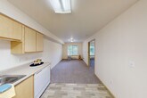 This one-bedroom, one-bathroom residence in Seattle's 98133 neighborhood offers a thoughtfully designed layout across a single floor. The kitchen is a functional highlight, featuring light maple cabinetry with ample upper and lower storage, a neutral countertop, and white appliances including a refrigerator, range, and dishwasher. Light wood-style flooring runs through the kitchen area, complemented by a skylight-style overhead fixture that keeps the space bright and welcoming. The kitchen opens directly into the living room, which is finished with plush carpet and benefits from natural light streaming through windows that frame views of the surrounding greenery — a serene nod to the Pacific Northwest setting. The bedroom is a comfortable, carpeted retreat with a window offering additional natural light and access to a closet. A second closet provides extra storage convenience. The bathroom features a wood-toned vanity with ample counter space, a large mirror with overhead lighting, and matching light flooring. Throughout the unit, warm neutral tones, light wood trim accents, and clean lines create a cohesive, understated aesthetic that is both practical and inviting.