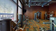 Located at 240 Fifth St E in Saint Paul, Minnesota, this fitness center offers a well-equipped, single-floor workout facility designed to support a wide range of exercise routines. The space is characterized by warm, earthy tones on the walls, complemented by a bold, multicolored patterned carpet that adds a lively visual energy throughout.

Large windows line the perimeter, flooding the interior with natural light and offering street-level views of surrounding trees and the urban neighborhood outside. Adjustable blinds provide control over lighting conditions during workouts.

The cardio section is outfitted with multiple treadmills and elliptical machines, including Precor XT4700 ellipticals, positioned to face the windows for an engaging workout experience. Wall-mounted flat-screen televisions are strategically placed for entertainment during cardio sessions.

The strength training area features a comprehensive selection of Paramount cable and selectorized machines, a multi-station cable system, and a full rack of dumbbells in varying weights. Large floor-to-ceiling mirrors expand the sense of space and allow for proper form checks during exercise.

Exposed ductwork on the ceiling contributes an industrial aesthetic, while recessed and track lighting ensures the space remains well-lit. The layout is thoughtfully organized to separate cardio and strength zones, providing an efficient and comfortable fitness environment.