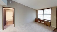 This single-floor space presents a clean, functional layout with a straightforward design. The main room is finished with wall-to-wall carpet in a neutral beige tone, providing a soft underfoot feel that complements the grey-painted walls. A large window on one wall allows natural light to fill the room, fitted with horizontal blinds for adjustable privacy and light control. Beneath the window sits a low wooden panel housing floor-level heating and ventilation units, a practical feature that contributes to efficient climate control throughout the space. The walls are smooth and minimally detailed, offering a blank canvas well-suited for personalization. An open doorway leads to an adjoining room, where hardwood or laminate flooring is visible, suggesting a transition to a different functional area — likely a hallway or bathroom, as indicated by the lighter interior beyond. The ceiling is standard height and smooth in finish. Overall, the space maintains a neutral, understated aesthetic with practical finishes, making it a versatile and livable environment ready for furnishing and personalization.