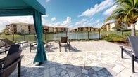 This outdoor amenity space is designed to deliver a resort-style experience, centered around a generously sized swimming pool surrounded by an expansive stone-paved deck. The deck is finished in a multi-toned, geometric paver pattern that adds visual texture and a polished aesthetic underfoot. Rows of dark lounge chairs line the poolside, providing ample space for sunbathing and relaxation under the open Florida sky.

A standout feature of this space is the collection of teal cabanas positioned along the deck, each outfitted with draped curtains, wicker seating, and round dining tables. These shaded retreats offer a more private and sheltered outdoor experience, ideal for small gatherings or leisurely afternoons. The teal color of the canopies adds a vibrant, tropical accent that complements the surrounding landscape.

At the perimeter of the paved area, a wrought iron fence borders a serene waterfront view, where a fountain-adorned lake reflects the surrounding multi-story buildings framed by palm trees. This lakeside backdrop adds a tranquil, scenic quality to the overall atmosphere. The combination of thoughtful design elements, lush tropical landscaping, and open water views creates a welcoming and visually cohesive outdoor retreat.