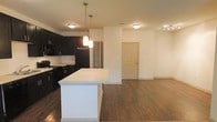 This single-floor residence offers a well-designed open-concept layout that connects the kitchen and living area in a cohesive, functional flow. The kitchen is a standout feature, showcasing rich dark cabinetry that contrasts beautifully with a clean white countertop island, providing ample prep and gathering space. A classic white subway tile backsplash adds a crisp, contemporary touch, while black appliances — including a refrigerator and range — complement the bold cabinetry. Pendant lighting hangs above the island, offering focused task lighting and a stylish design accent. The open layout extends seamlessly into the living area, which is finished with warm hardwood-style flooring that runs throughout the space, adding texture and visual continuity. The neutral white walls and flush-mount ceiling lights keep the interior feeling bright and airy. A private entry door is visible at the far end of the living area, offering a defined sense of arrival. Overall, the space combines modern aesthetics with practical design, making it an inviting and versatile environment suited for both everyday living and entertaining.