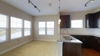 This single-floor space showcases an open-concept layout that seamlessly connects the kitchen and living area, creating a cohesive and functional environment. The kitchen is outfitted with rich dark wood cabinetry that provides a striking contrast against the light-toned walls. A granite countertop peninsula with an undermount stainless steel sink serves as a natural divider between the two areas, while a pendant light above adds a refined touch. Additional upper cabinetry offers ample storage.

The living area features plush carpeting in a neutral tone, contributing to a warm and comfortable atmosphere. Track lighting along the ceiling provides adjustable, directed illumination throughout the space. Multiple windows fitted with horizontal blinds line the walls, inviting generous natural light and offering pleasant views of the surrounding outdoor scenery, including what appears to be a waterfront or open landscape. The combination of modern fixtures, cohesive color palette, and open sightlines gives this space a clean, contemporary feel well-suited for everyday living.