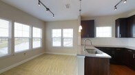This single-floor space showcases an open-concept layout that seamlessly connects the kitchen and living area, creating a cohesive and functional environment. The kitchen is outfitted with rich dark wood cabinetry that provides a striking contrast against the light-toned walls. A granite countertop peninsula with an undermount stainless steel sink serves as a natural divider between the two areas, while a pendant light above adds a refined touch. Additional upper cabinetry offers ample storage.

The living area features plush carpeting in a neutral tone, contributing to a warm and comfortable atmosphere. Track lighting along the ceiling provides adjustable, directed illumination throughout the space. Multiple windows fitted with horizontal blinds line the walls, inviting generous natural light and offering pleasant views of the surrounding outdoor scenery, including what appears to be a waterfront or open landscape. The combination of modern fixtures, cohesive color palette, and open sightlines gives this space a clean, contemporary feel well-suited for everyday living.